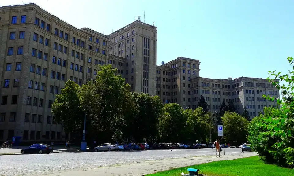 Великий ступінчастий фасад головного корпусу Каразінського університету серед зелених дерев під ясним небом; унизу кілька людей і автомобілів