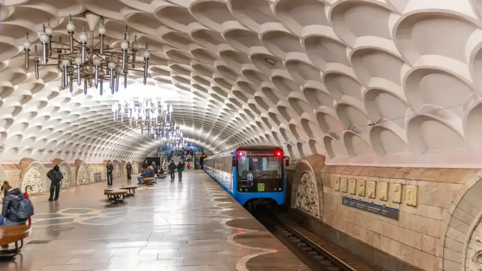Просторий перон станції метро «Київська» зі світлою рельєфною стелею, синім потягом, назвою станції на стіні та кількома пасажирами