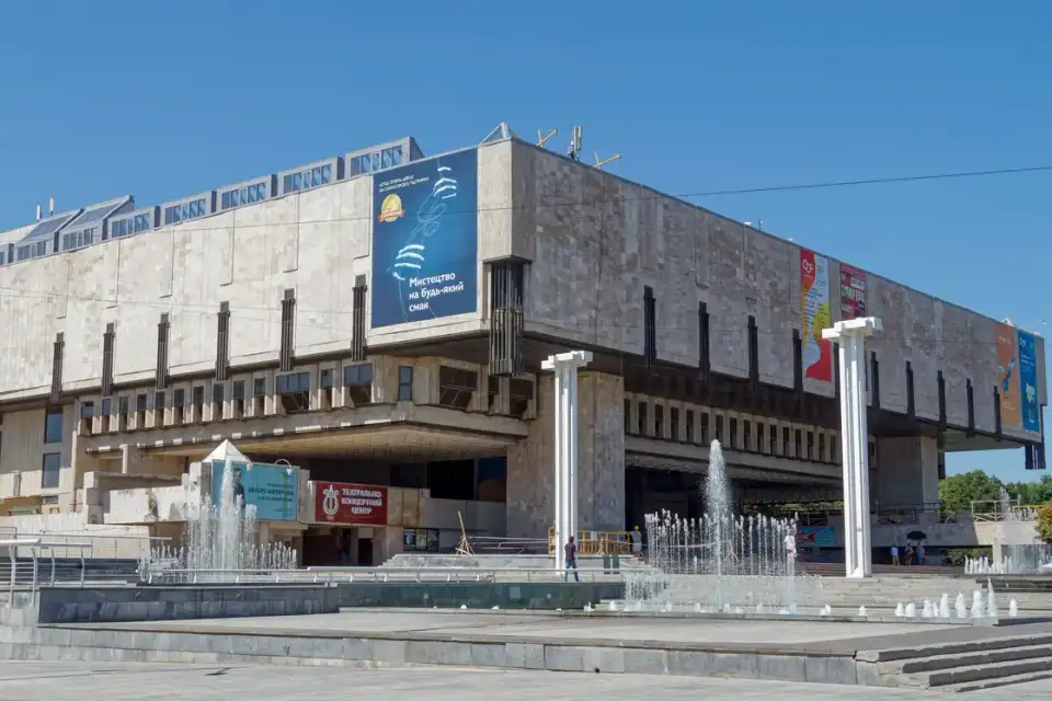Великий світлий фасад Харківської національної опери за двома фонтанами під ясним небом; перед будівлею видно лише кількох людей
