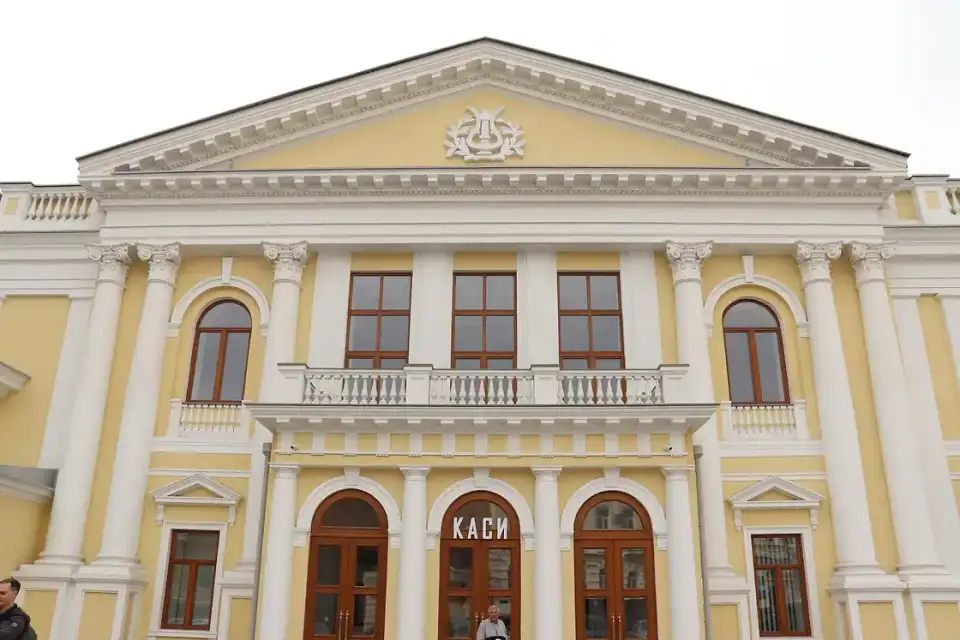 Світло-жовтий симетричний фасад філармонії з білими колонами, балконом і написом Каси над центральним входом; унизу двоє людей