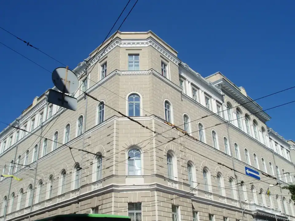 Світло-сірий кутовий фасад Будинку Держбанку з рядами аркових вікон під чистим синім небом; перед верхніми ярусами проходять тролейбусні дроти