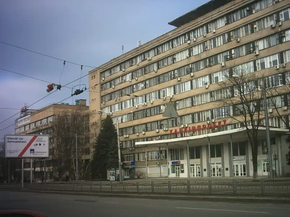 Протяжний цегляний фасад із червоним написом «Харківпроєкт», порожнім тротуаром, огорожею та безлистими деревами