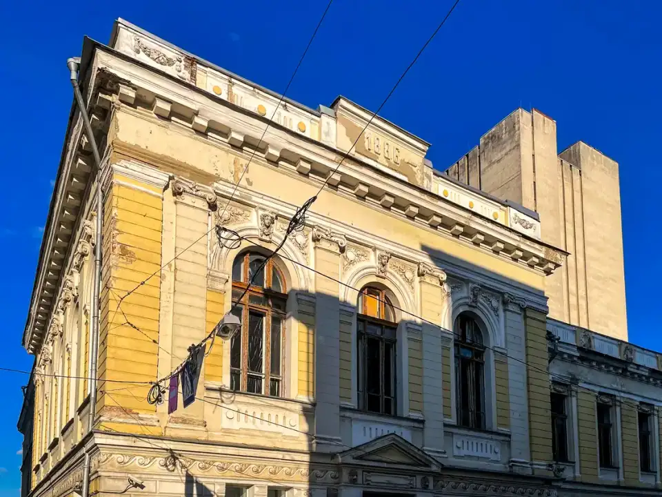 Історичний жовто-білий фасад бібліотеки Короленка під синім літнім небом; на карнизах і оздобленні видно сліди пошкодження, людей у кадрі немає