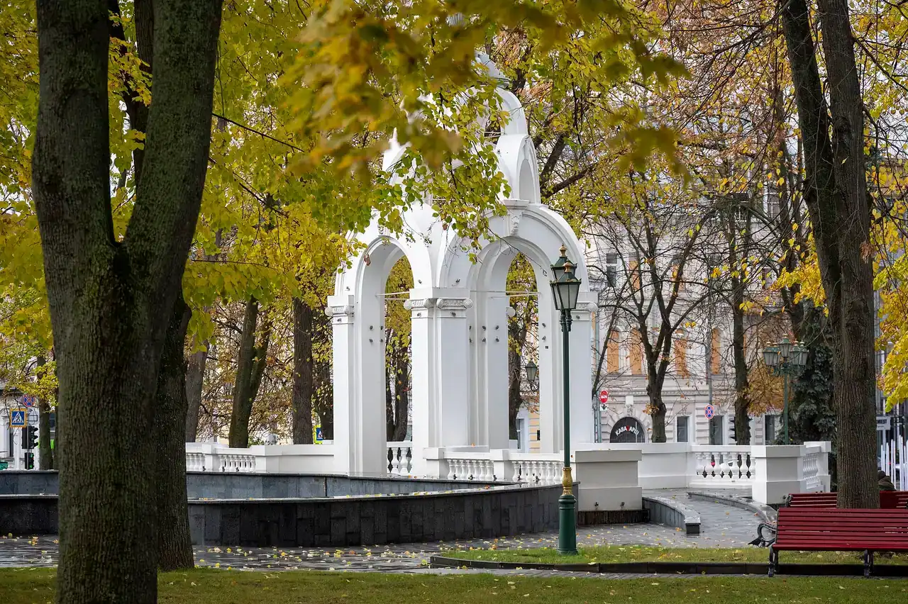 Біла аркова альтанка Дзеркального струменя між жовтим листям, деревами та лавами