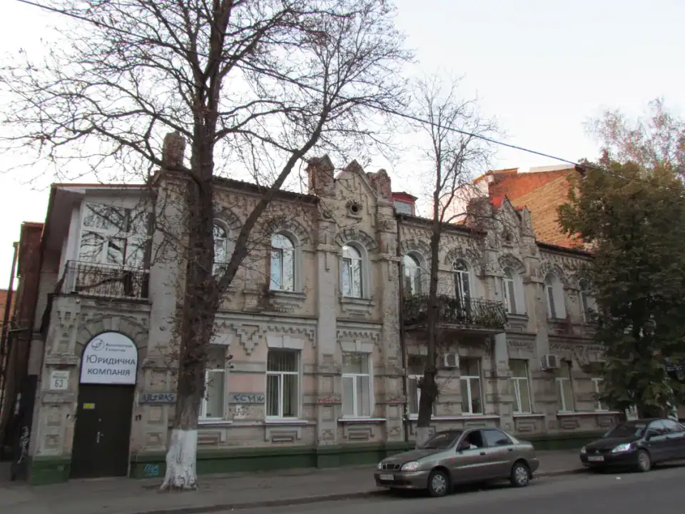 Двоповерховий цегляний фасад із арковими вікнами, балконами, деревами, номером 63 біля лівих дверей і без людей на тротуарі