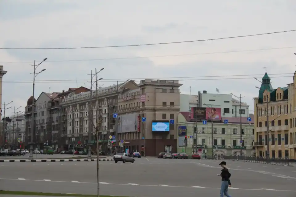 Просторий Павлівський майдан із рядами різностильових фасадів, кількома автомобілями та однією перехожою на передньому плані