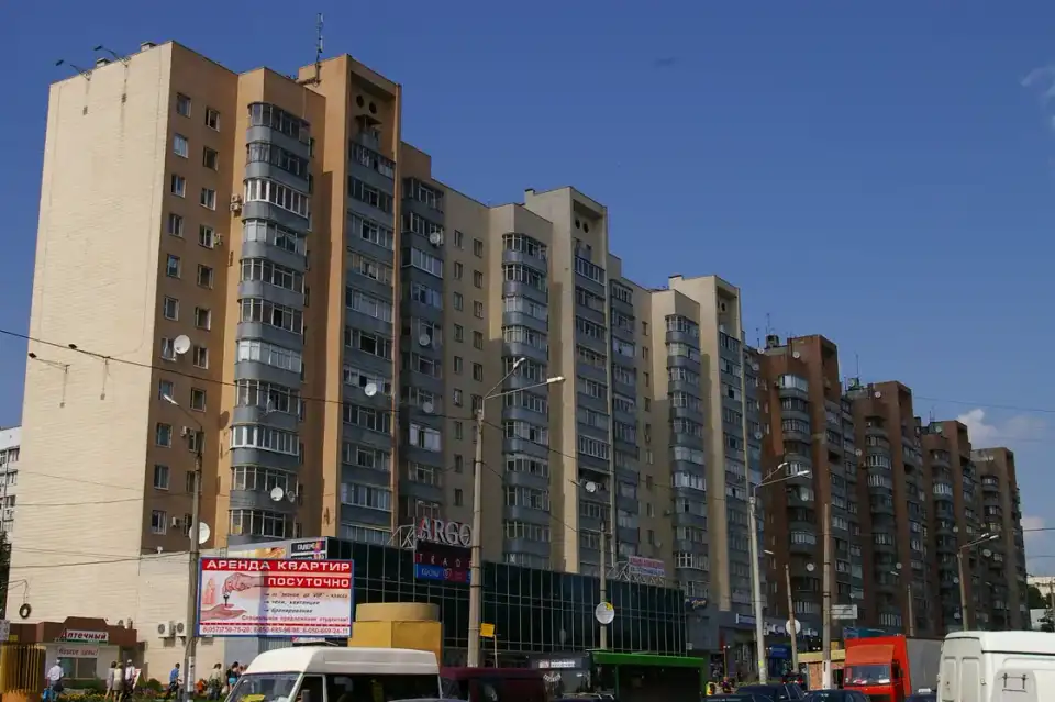Довгий багатоповерховий фасад на Полтавському Шляху під синім літнім небом, із транспортом та кількома людьми внизу кадру