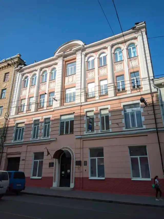 Рожево-білий фасад Державного архіву Харківської області з арковим входом, настінними табличками та прапором; біля краю кадру стоять один перехожий і фургон