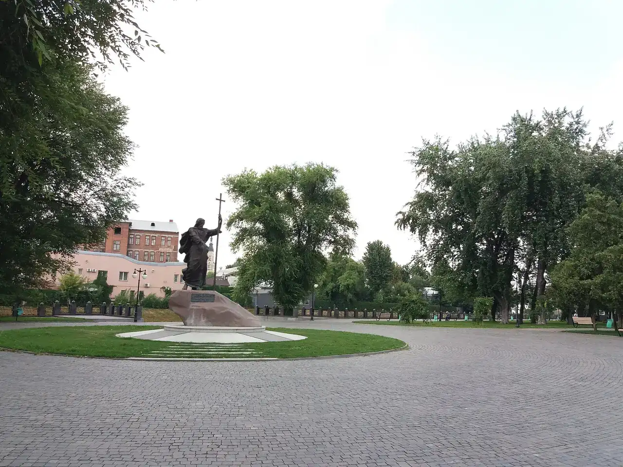 Простора вимощена алея скверу з деревами, лавами й памʼятною скульптурою на газоні