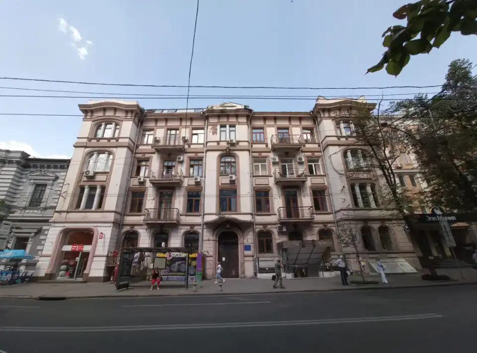 Світлий чотириповерховий фасад на Сумській із балконами, арковими вікнами, зеленим деревом, зупинкою та кількома людьми на тротуарі