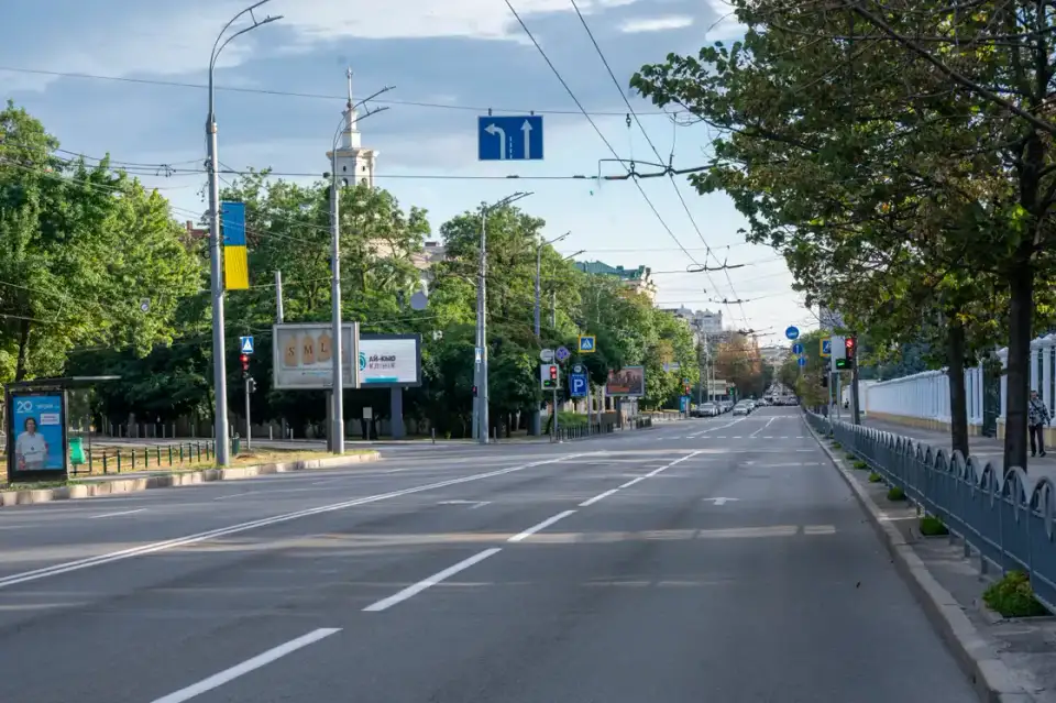 Майже порожня широка Сумська вулиця влітку з зеленими деревами, тролейбусними дротами, дорожньою розміткою та двома людьми вдалині