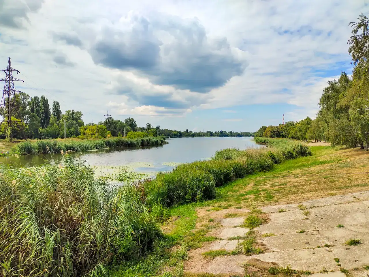 Широка водна гладінь Удянського гідропарку з очеретом, зеленими берегами й хмарним літнім небом без людей у кадрі