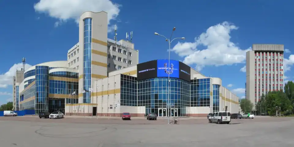 Широкий світлий фасад комплексу UNIT.Kharkiv зі скляними секціями, синьою вивіскою, майже порожньою площею та зеленими деревами